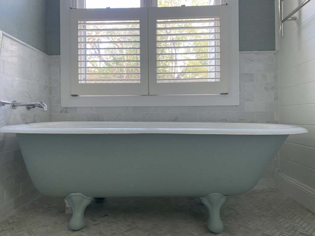 Bathroom with freestanding bathtub, green wallpaper above tiled walls, white shutters, and natural light through window