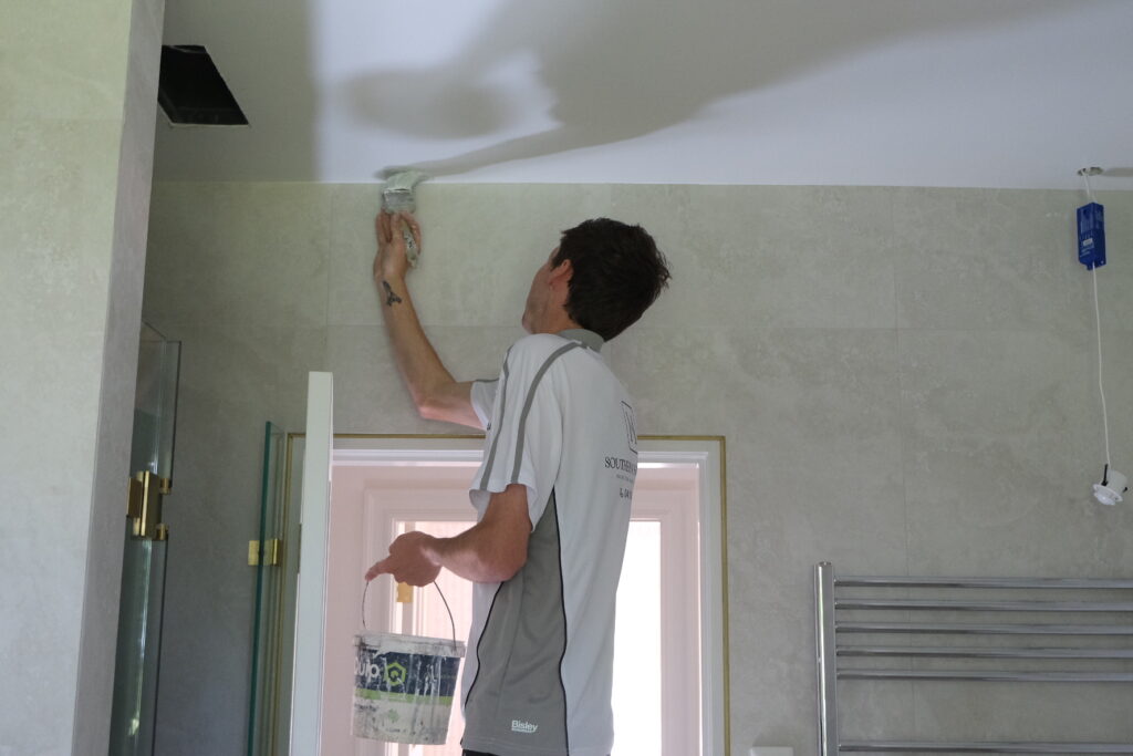 A worker from Southern Highlands Painting and Wallpapering paints a bathroom ceiling. The room has tiled walls, a towel rack, and a glass shower door with gold trim.