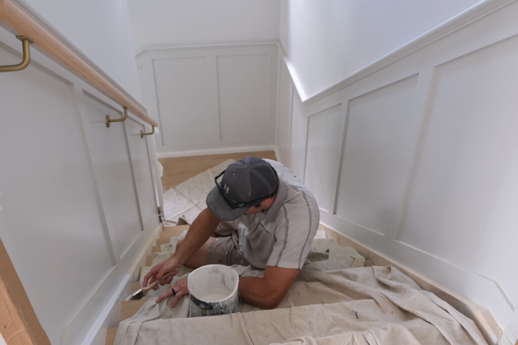 A worker from Southern Highlands Painting and Wallpapering paints the staircase trim, surrounded by white wainscoting, a wooden handrail, and a protective drop cloth.