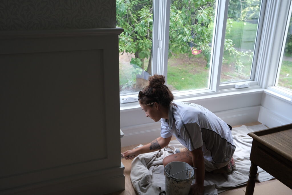 A worker from Southern Highlands Painting and Wallpapering paints a white baseboard near a large window, with a drop cloth protecting the wooden floor.