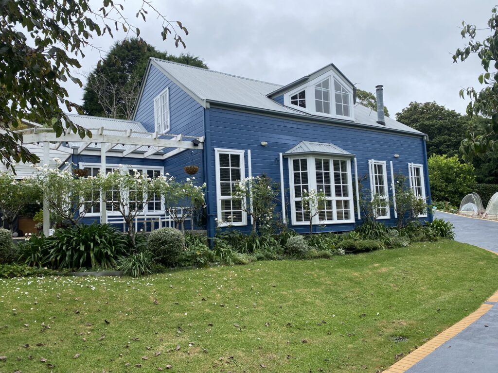 Charming two-storey weatherboard home painted in coastal blue with white trim, surrounded by lush garden and rose bushes, Southern Highlands NSW.