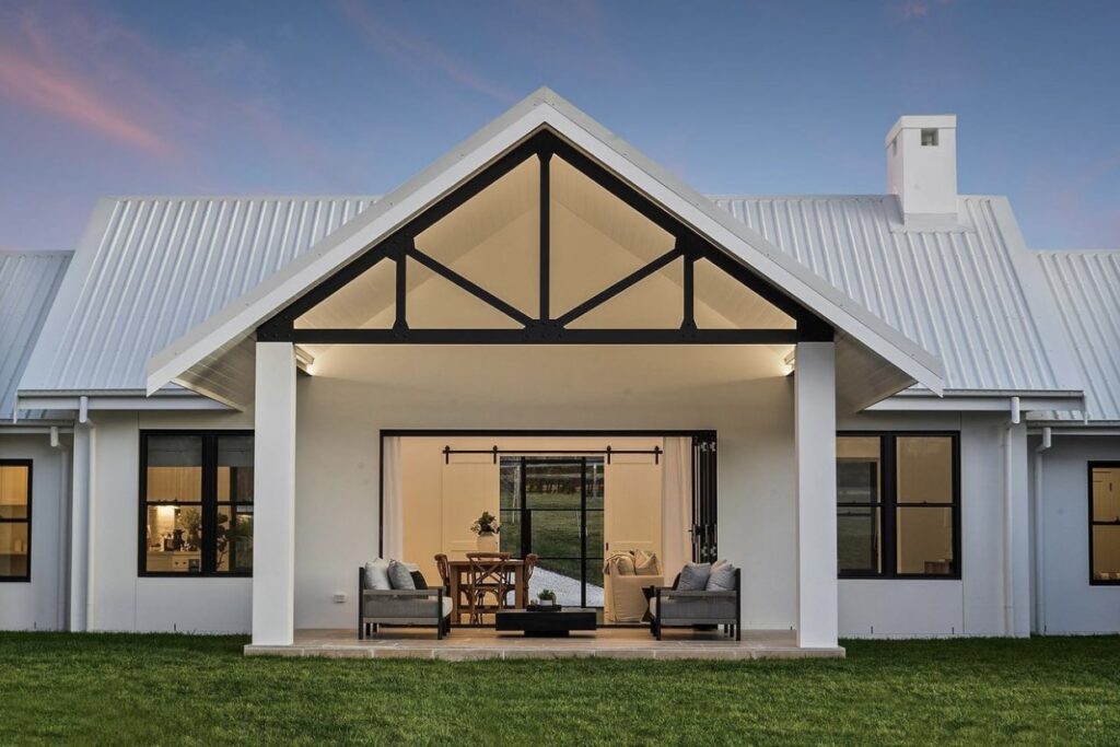 Contemporary white farmhouse with pitched metal roof, black window frames, open gable patio, and inviting alfresco area at sunset.
