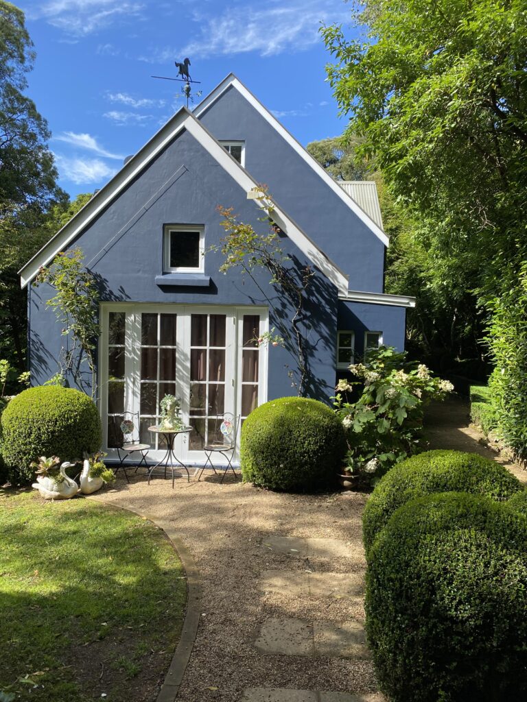 Charming blue cottage with white trim nestled in a lush garden, featuring manicured hedges, gravel pathway, French doors, and decorative swan statues under a clear blue sky.