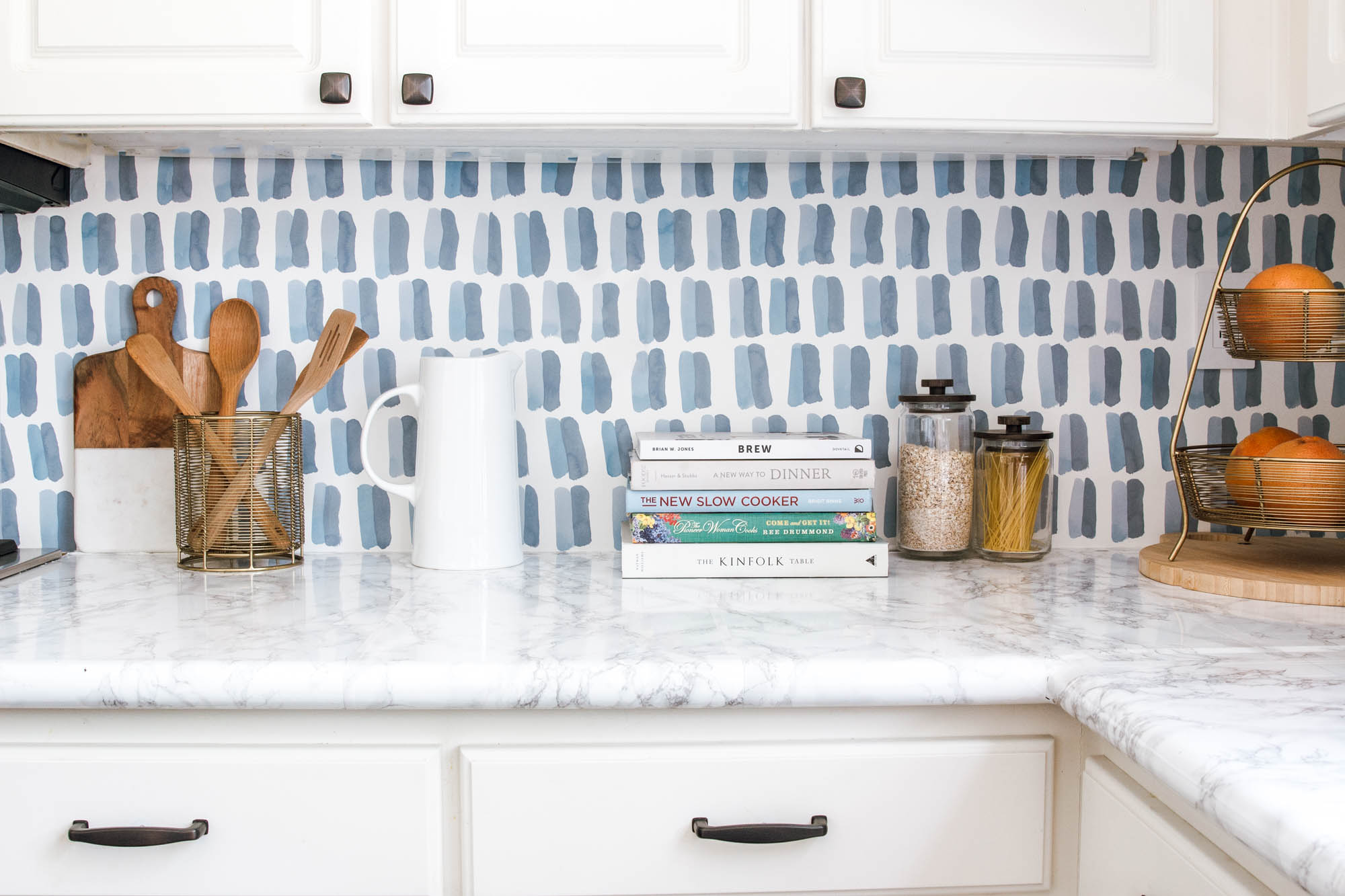 Kitchen with blue patterned wallpaper splashback, white cabinetry, marble benchtop and styled cooking accessories