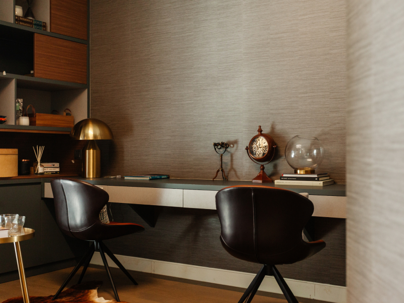 Home office with textured neutral wallpaper, built-in desk, dark chairs and warm ambient lighting